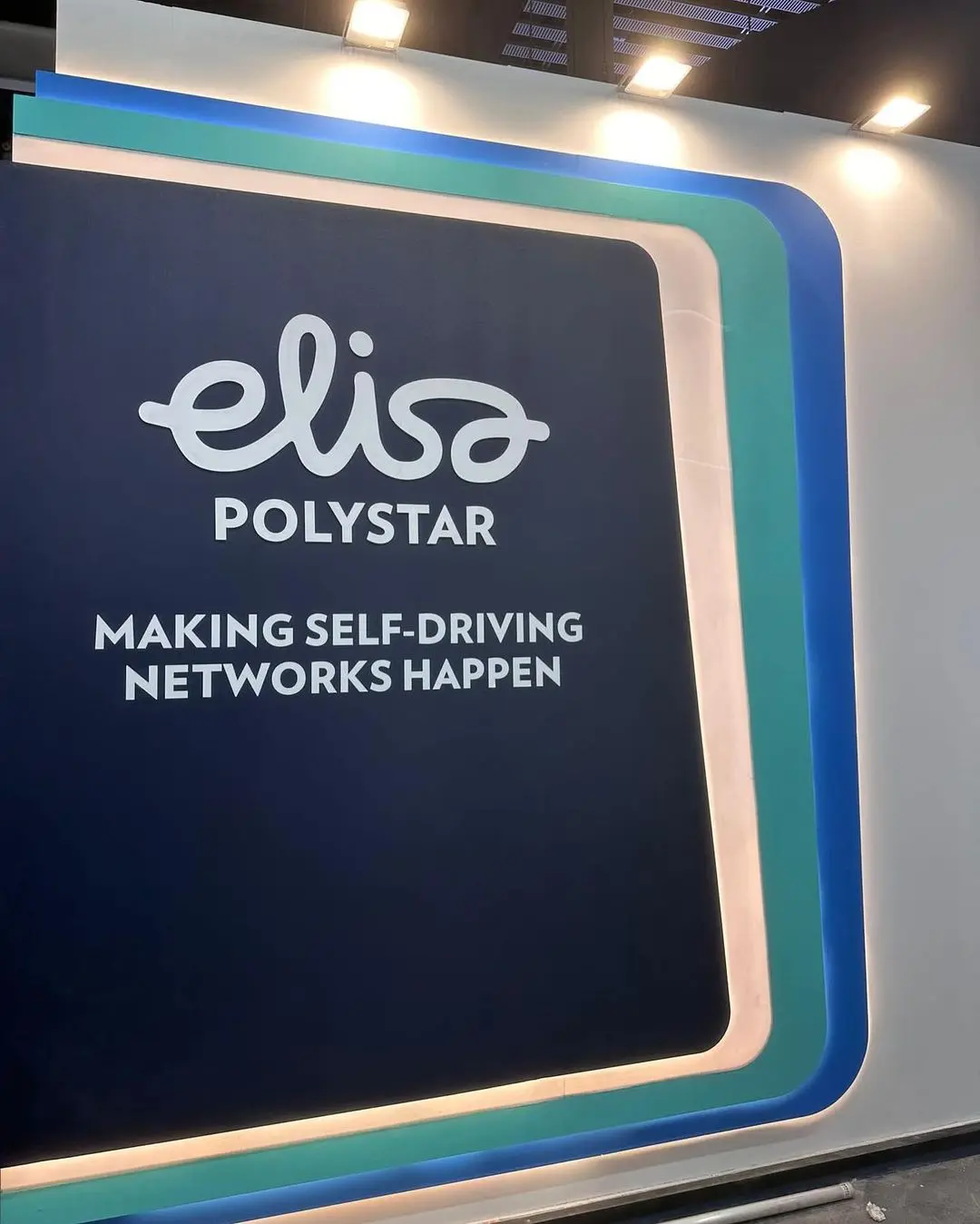 Un stand de Elisa Polystar en una feria comercial. El letrero presenta el logotipo de la empresa y el lema "Making Self-Driving Networks Happen" sobre un fondo azul marino con detalles en verde y verde azulado.