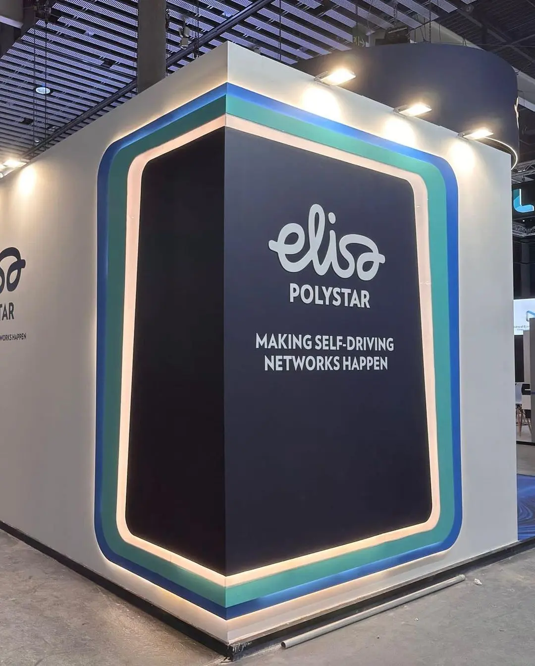 Un stand de feria comercial con un fondo azul oscuro presenta el logotipo y el nombre "Elisa Polystar" en texto blanco. El lema "Haciendo realidad las redes autónomas" se muestra debajo del nombre. El stand es moderno con detalles de iluminación geométricos en los bordes.