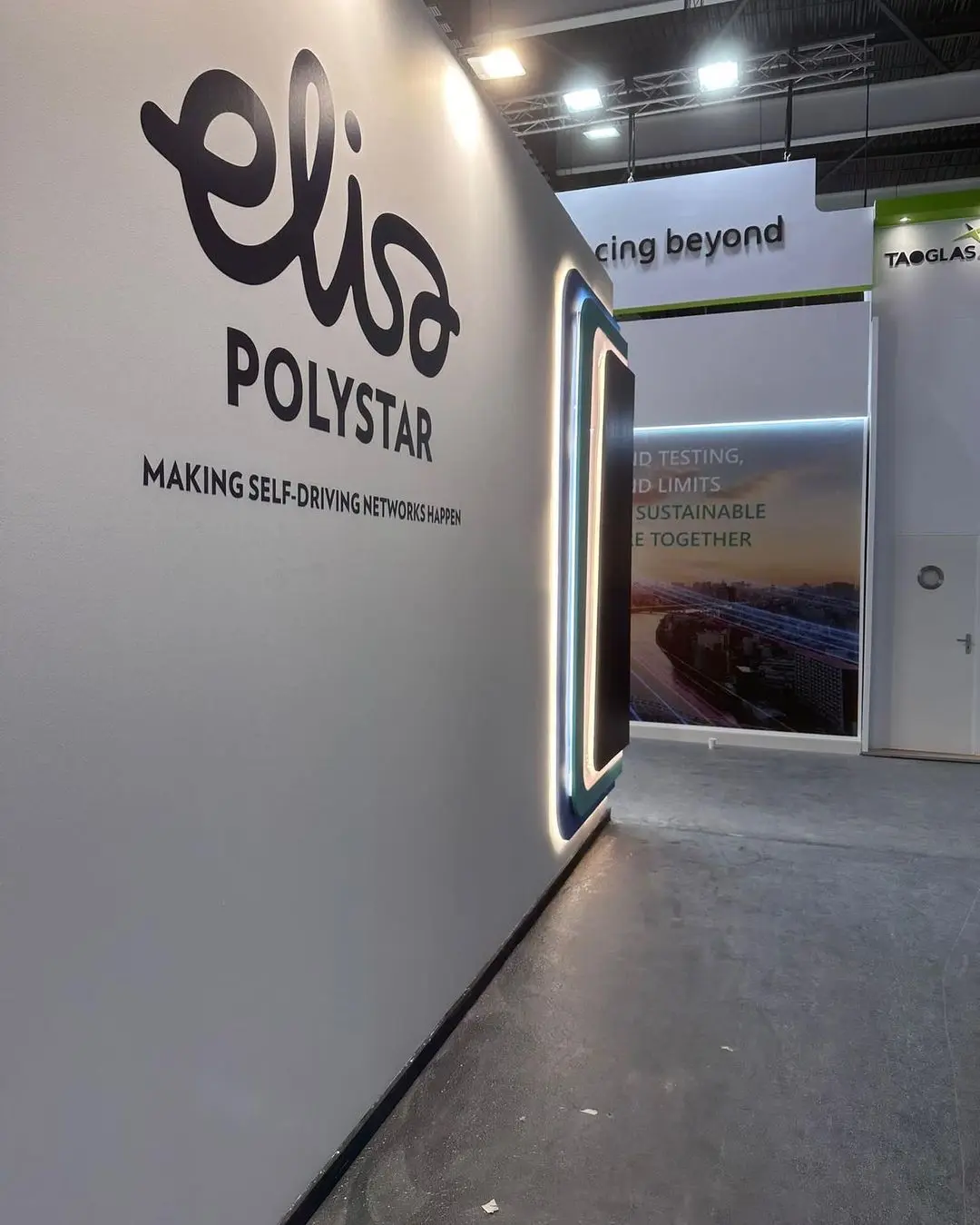 Stand en una exposición que presenta a Elisa Polystar. Un letrero que dice "Elisa Polystar haciendo realidad las redes autónomas" se exhibe de manera destacada en una pared con bordes iluminados. Al fondo, un pasillo conduce a otra zona expositiva con diversos carteles y pancartas.