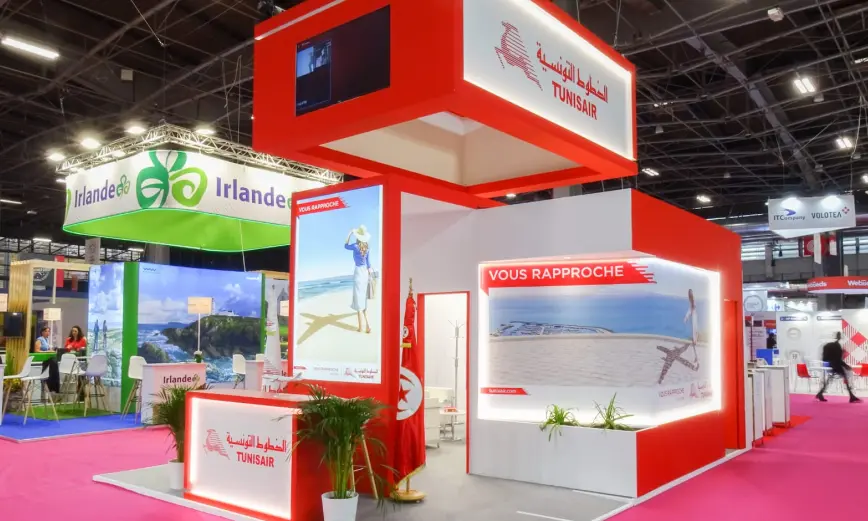 Un stand de feria luminoso para Tunisair con colores rojo y blanco. El stand incluye un mostrador de recepción, grandes gráficos de escenas de viajes y pancartas con el logotipo de Tunisair. Hay carteles de otros stands como Irlandee visibles al fondo.