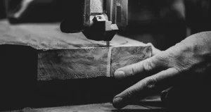 Primer plano en blanco y negro de una escena de carpintería, que muestra la mano de una persona guiando un trozo de madera a través de una sierra de cinta. La imagen detallada captura la textura de la madera y la precisión de la hoja de sierra en movimiento.