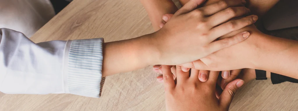 Un grupo de personas coloca sus manos una encima de la otra en una muestra de unidad y trabajo en equipo. Las manos son de diferentes tamaños y tonos de piel, lo que significa diversidad. El fondo es una mesa de madera clara.