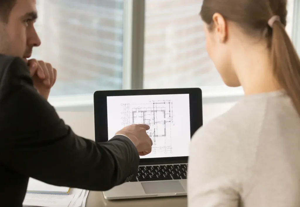 Dos personas sentadas en un escritorio, examinando un plano mostrado en la pantalla de una computadora portátil. La persona de la izquierda apunta a la pantalla y la persona de la derecha está de espaldas a la cámara. Entorno de oficina con ventanas al fondo.